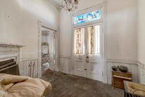 A worn, vintage entryway with high ceilings, stained glass windows above double doors, peeling white wood paneling, a fireplace, and various household items scattered throughout. A patterned room is visible through the doorway.