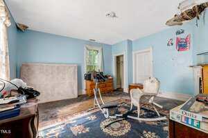 A cluttered room with blue walls, a wicker chair, an exercise bike, a mattress propped against the wall, a dresser, a small fireplace, peeling paint on the ceiling, and a Ouija board on a nearby table.