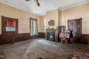 A mostly empty, old-fashioned room with wood trim, a fireplace with a mirror above it, a small table and chair, dusty wooden floors, and some items scattered on the floor, including an orange extension cord.