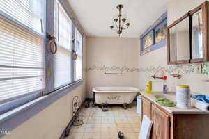 A bathroom with a clawfoot bathtub, peeling floral wall border, worn linoleum floor, and a wooden vanity with cleaning supplies. Three large windows with blinds line one wall, and a vintage light fixture hangs from the ceiling.
