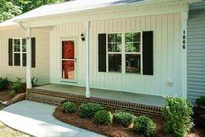 A single-story house with white siding, black shutters, a red front door, and brick steps leading to a covered front porch. The entryway is bordered by shrubs and mulch. House number 1006 is visible.