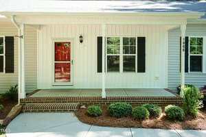 A white house exterior with black shutters, a red front door with a glass storm door, a covered porch with brick steps, small shrubs, and house number 1006 displayed vertically on the wall.