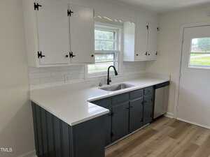 A modern kitchen with white upper cabinets, dark gray lower cabinets, a white countertop, white tile backsplash, stainless steel sink with black faucet, dishwasher, and a window above the sink.