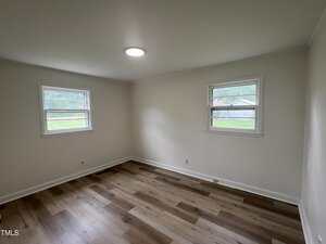 Empty room with light-colored walls, two windows on adjacent walls, ceiling light fixture, and wood-style flooring. Natural light enters through the windows, showing a green yard and fence outside.