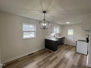 A kitchen with light wood floors, white walls, black cabinets, white countertops, a farmhouse sink, an oven, and a large black light fixture hanging from the ceiling. Two windows and a back door provide natural light.