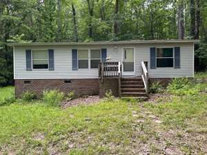 A single-story house with white siding, brick foundation, blue shutters, and a small front porch with steps, nestled in a wooded area with overgrown grass at 3539 Charlies Court in Wake Forest.