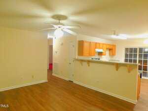 A well-lit open-concept room with wood flooring, beige walls, a ceiling fan, and a kitchen area featuring light wood cabinets and a breakfast bar; a sliding glass door is visible in the background.