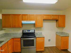 A kitchen with wood cabinets, granite countertops, stainless steel electric stove, range hood, and an empty space likely for a refrigerator; there is also a laundry hookup on the wall and wood-style flooring.