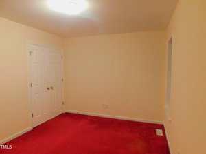 A small, empty room with cream-colored walls, a red carpet, a white double door, a ceiling light, and a rectangular window or wall cutout on the right. Electrical outlets and a floor vent are visible.