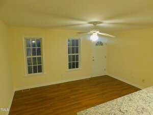 A room with light yellow walls, two tall windows, a white door, a ceiling fan with a light, and wood flooring. The edge of a granite countertop is visible in the lower right corner.
