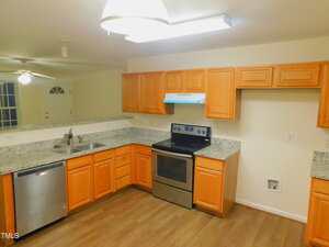 A kitchen with light wood cabinets, granite countertops, stainless steel appliances including a stove and dishwasher, a double sink, and wood flooring. An open pass-through connects to the adjacent room.