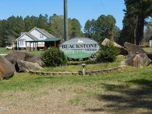 A large oval sign reading "Blackstone Village" sits among bushes and rocks on a grassy area near 1098 Blackstone Drive, with houses and trees in the background under a clear Creedmoor sky.