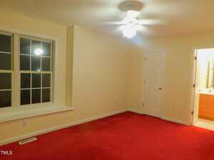 A room with cream-colored walls, a red carpet, a ceiling fan with a light, a window with white trim, a white door, and an open door leading to a bathroom with a visible vanity and mirror.