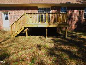 A wooden deck with stairs attached to the back of a red brick house, featuring two windows and a glass door. The deck is elevated with support posts and casts a shadow on the ground covered in dry leaves.