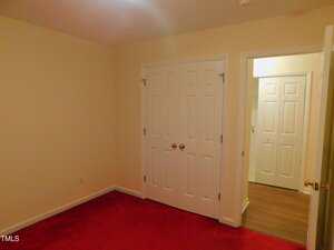 A small room with beige walls, a red carpet, a closed white double closet door, and an open door leading to a hallway with wood flooring and another white door.