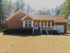 A single-story brick house with a front porch, white railings, a set of steps leading to the entrance, and an attached garage. The yard is mostly dry grass with trees in the background.