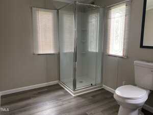 A bathroom with a corner glass shower, a white toilet, two windows with blinds, light-colored walls, and wood-look flooring.