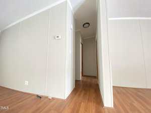 A hallway with light-colored walls and wood laminate flooring, featuring a visible thermostat, an overhead light, and a partially visible corner of an adjacent room.