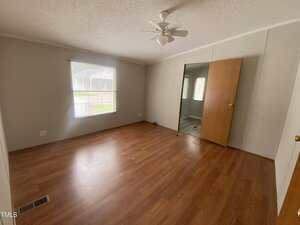 An empty room with wood flooring, light-colored walls, a ceiling fan, a large window with blinds, and two wooden doors, one partially open showing an adjacent room. Natural light comes in through the window.