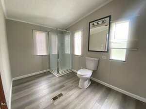 A bathroom with light gray walls and flooring, featuring a glass shower enclosure, a toilet, a large wall mirror, and three windows with blinds that let in natural light.