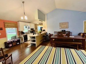 Open-concept kitchen and dining area with white cabinets, stainless steel appliances, a large wooden dining table, several chairs, and a striped area rug on dark hardwood floors. Walls are painted yellow and light blue.