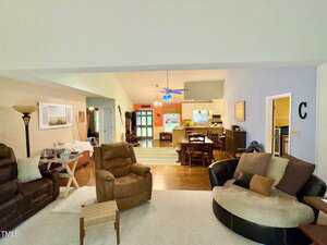 Spacious open-concept living and dining area with beige walls, various chairs and sofas, a round chair with pillows, wooden dining table, kitchen in the background, and ceiling fan with blue lights.