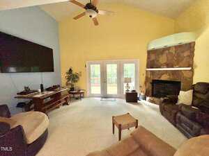 A living room with beige carpet, yellow and blue walls, a mounted TV, a round chair, brown couches, a wooden table, French doors leading outside, and a stone fireplace in the corner. A ceiling fan hangs above.