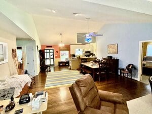 Open-concept living space with wood floors, a dining area, and kitchen. Brown recliner in foreground, striped rug, ceiling fan with blue light, and various furniture. Doorways lead to other rooms. Walls are light blue and cream.
