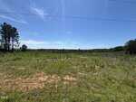 A wide, open field with sparse grass and patches of bare soil under a clear blue sky sits near 00000 Cheeks Quarter Road in Henderson. Trees line the edges, with a few small stumps and scattered debris visible in the center.