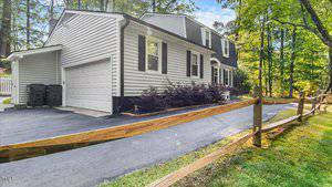 A two-story house with gray siding, black shutters, a white garage door, and a paved driveway. There is a wooden fence bordering the driveway and a backdrop of tall trees and greenery.