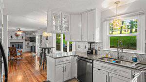 A modern kitchen with white cabinets, stainless steel appliances, granite countertops, and a double sink under a window. The space opens to a living area with a fireplace, wood floors, and large windows.