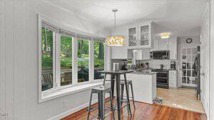 A kitchen with white cabinets, stainless steel appliances, a small bar table with two stools, hardwood floors, and a large bay window overlooking a green backyard. Pendant lights hang from the ceiling.