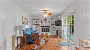 A living room with white walls and wood floors features a blue sofa, wooden chair, ceiling fan, fireplace with shelves, wall-mounted TV, and sliding glass doors leading to a deck with greenery outside.