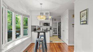 Bright kitchen with white cabinets, stainless steel appliances, a peninsula with barstools, wood floors, and large windows overlooking greenery; framed art hangs on a light gray wall.