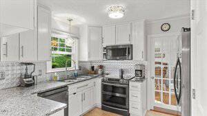 A modern kitchen with white cabinets, stainless steel appliances, granite countertops, and a metallic tile backsplash. There is a window above the sink and a glass-paneled door near the refrigerator.