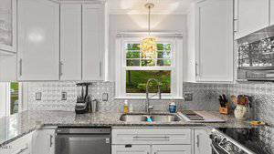 A modern kitchen with white cabinets, granite countertops, a double sink under a window, stainless steel dishwasher, blender, soap dispensers, and various utensils on the counter. Patterned tile backsplash is visible.