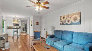 A living room with wood flooring, a blue couch, a wooden chair, a coffee table, and a painting of dandelions on the wall. The room connects to a kitchen with bar stools and white cabinets. A ceiling fan with lights is on the ceiling.