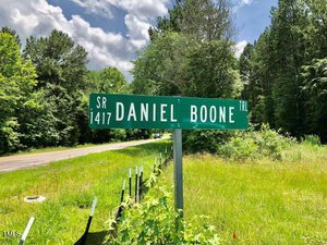 A green street sign with white letters reads "SR 1417 Daniel Boone Trl" beside a rural road, surrounded by tall grass, trees, and a partly cloudy sky.