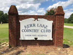 A large white sign with black text reads "Kerr Lake Country Club Green Fees Accepted," mounted on a brick structure outdoors with grass and trees in the background.