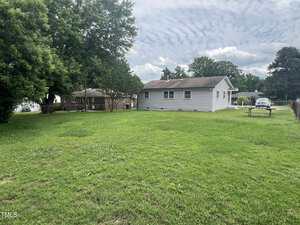 Single-story house with white siding sits on a large grassy backyard. A few trees and a wooden picnic table are visible, with another house and several cars in the background. Sky is mostly cloudy.