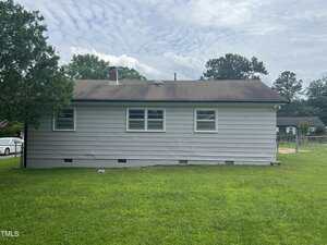 A single-story house with light gray horizontal siding, a dark shingled roof, and three windows, seen from the backyard with green grass and a partially visible tree to the left.