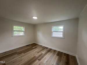 Empty room with light-colored walls, two windows showing greenery outside, a ceiling light, and wood-patterned laminate flooring. The space is unfurnished and appears clean and newly renovated.