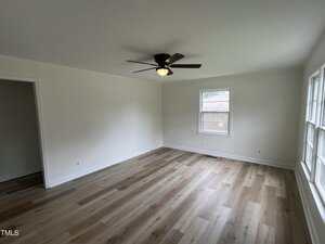 A bright, empty room with light wood flooring, white walls, a black ceiling fan with a light, a single window on one wall, and multiple large windows on another wall. An open doorway leads to an adjacent space.