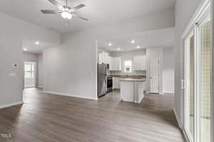 Open-concept living area with light wood flooring, a ceiling fan, and sliding glass doors leading outside. The modern kitchen features white cabinets, stainless steel appliances, and recessed lighting.