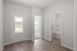 A hallway with light wood flooring, white walls, a frosted glass door on the left, and two open doorways leading to a bedroom and a bathroom with a bathtub and shelves.