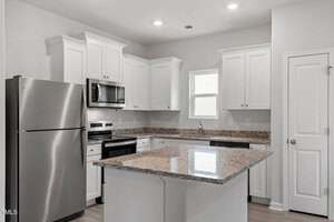 A modern kitchen with white cabinets, stainless steel appliances, a granite countertop island, single window above the sink, and a closed pantry door on the right. The room has neutral walls and recessed lighting.