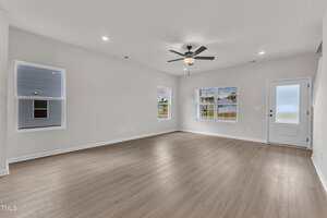 An empty living room with light gray walls, wood-look flooring, three large windows, a ceiling fan with a light, and a white door with frosted glass. Recessed ceiling lights are turned on.