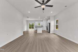 A spacious, empty open-concept living area with light wood floors, white walls, recessed lighting, ceiling fan, and a view of a modern kitchen with an island and stainless steel appliances.