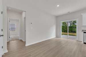 Unfurnished room with light wood flooring, white walls, and a sliding glass door leading outside. An open doorway on the left leads to another room, and there is a small hallway visible. Natural light enters through the windows.