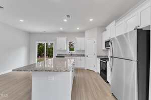 Modern kitchen with white cabinets, stainless steel appliances, granite countertops, a large island, light wood flooring, and recessed lighting. Sliding glass doors lead to an outdoor area with trees visible.